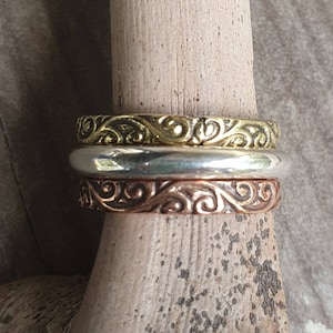 Peut inclure: Trois bagues métalliques empilées sur une cheville en bois. La bague supérieure est dorée avec un motif tourbillonnant orné. La bague du milieu est argentée et la bague inférieure est cuivrée, également avec un motif tourbillonnant. Les bagues sont probablement des bijoux.