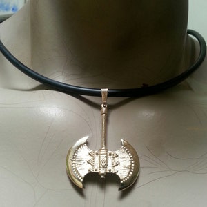 May include: A gold-colored pendant necklace featuring a double-bladed axe design. The axe pendant is suspended from a black cord necklace. The axe has detailed engravings and a polished finish, creating a striking visual contrast.