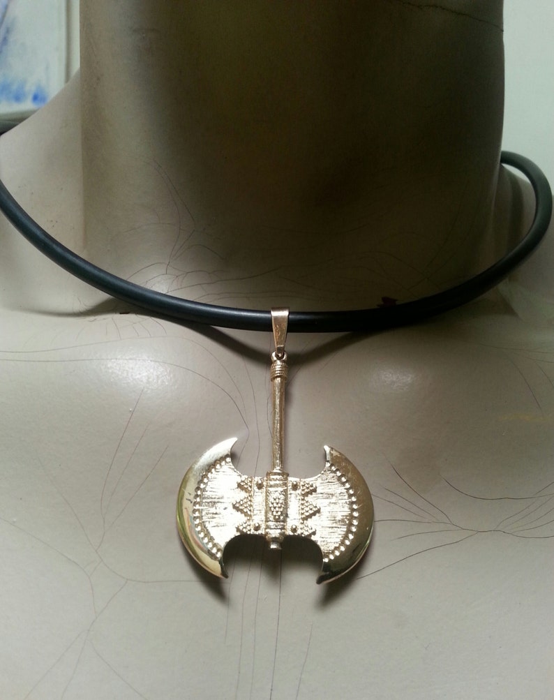 May include: A gold-colored pendant necklace featuring a double-bladed axe design. The axe pendant is suspended from a black cord necklace. The axe has detailed engravings and a polished finish, creating a striking visual contrast.
