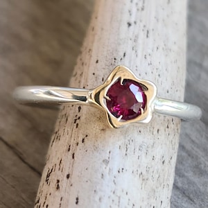 May include: A silver ring with a gold-coloured floral setting holding a round, deep red gemstone. The ring is displayed on a light-coloured wooden surface, showcasing the contrast between the metal and the stone.