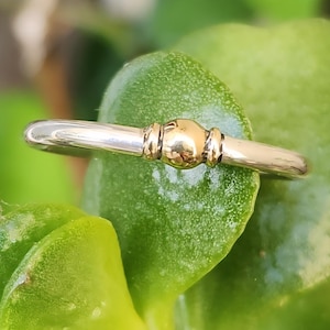 May include: A polished gold ring featuring a smooth, rounded band with a central spherical bead accent flanked by two small, vertical grooved details. The metal has a reflective, high-shine finish that catches the light, creating a bright appearance.