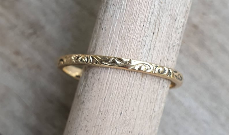 May include: A gold ring with an intricate floral pattern. The ring is a thin band, and the design wraps around the entire circumference. The ring is displayed on a light-colored wooden dowel, showcasing its details.