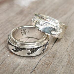 Könnte beinhalten: Zwei silberne Ringe mit Wellenmustern. Ein Ring ist auf dem anderen gestapelt. Die Ringe haben ein strukturiertes Wellenmuster und eine glatte, glänzende Oberfläche. Die Ringe liegen auf einer Holzoberfläche.