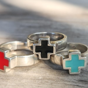 Könnte beinhalten: Drei silberne Ringe mit Kreuzdesigns in Rot, Schwarz und Türkis. Die Ringe werden auf einer Holzoberfläche präsentiert. Die Ringe sind ein Modeaccessoire.