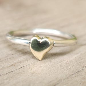 May include: A silver ring with a gold heart-shaped centerpiece. The ring is set against a light-colored wooden surface. The heart is smooth and polished, reflecting light.