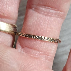May include: A gold-colored ring with an intricate, repeating swirl pattern. The ring is thin and delicate, with a polished finish. The design is visible on the outer surface of the ring. The ring is shown on a finger.