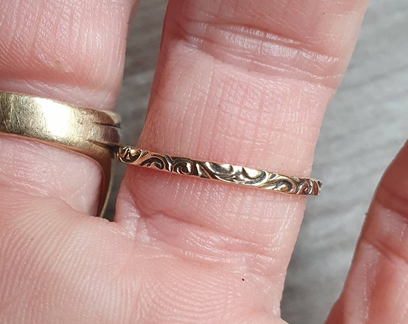May include: A gold-colored ring with an intricate, repeating swirl pattern. The ring is thin and delicate, with a polished finish. The design is visible on the outer surface of the ring. The ring is shown on a finger.
