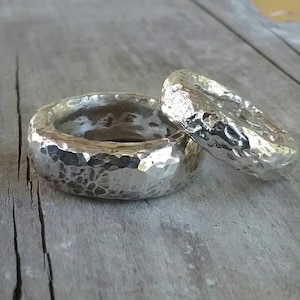 Puede incluir: Dos anillos de plata con textura martillada. Un anillo es más ancho que el otro. Los anillos se muestran sobre una superficie de madera envejecida. Los anillos tienen una superficie texturizada e irregular.