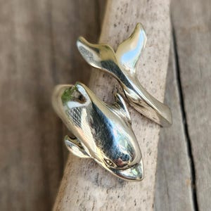 May include: A silver-colored ring in the shape of a dolphin, with the dolphin's body and tail forming the ring's design. The ring is shown on a piece of wood with a natural, light brown color.