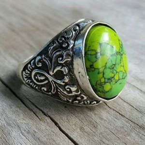 May include: A silver ring featuring an oval, lime green stone. The ring's band is decorated with an ornate, engraved pattern. The stone displays a mottled pattern of lighter and darker green shades, set in a silver bezel.