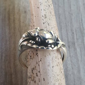 May include: A silver ring featuring a detailed frog design. The frog is resting on a lily pad, with intricate detailing on the ring's band. The ring is displayed on a wooden dowel, showcasing its design and craftsmanship.