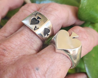 Puede incluir: Dos anillos de plata. Un anillo presenta el diseño de la carta As de Picas, con el símbolo de picas y la letra "A" en la cara. El otro anillo tiene forma de pica. Ambos anillos están en dedos.