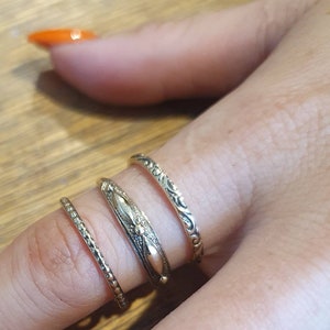 May include: Four gold-toned rings of varying designs are displayed on a finger. The rings include a textured band, a band with a central design, and two bands with ornate patterns. The rings are likely for fashion or jewelry.