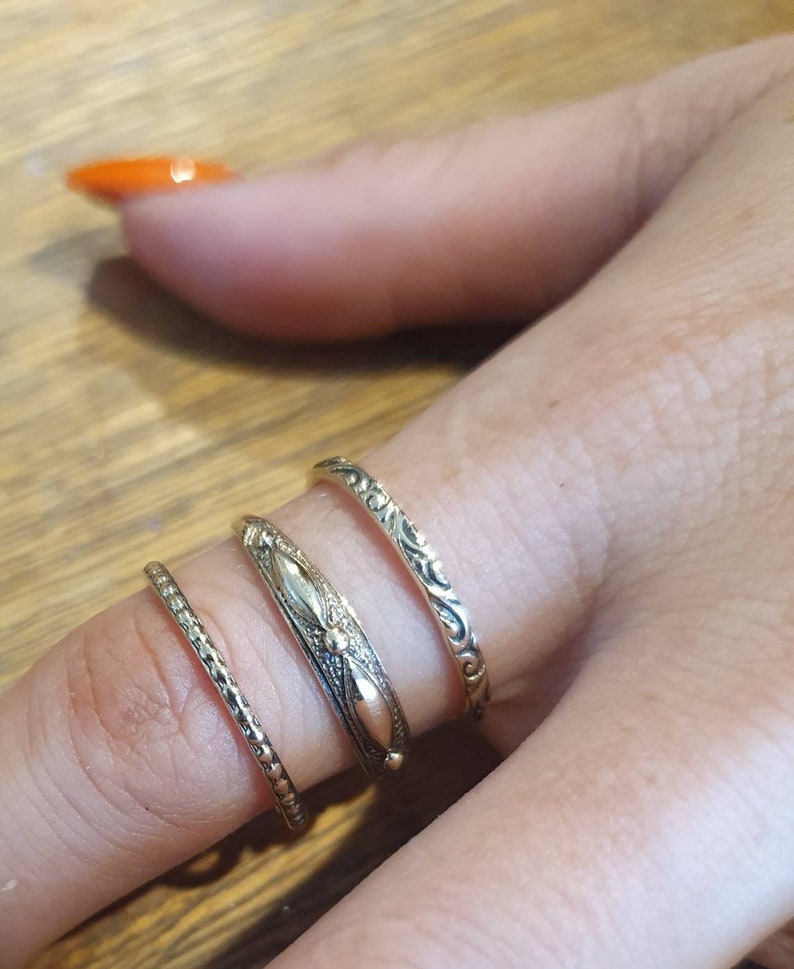 May include: Four gold-toned rings of varying designs are displayed on a finger. The rings include a textured band, a band with a central design, and two bands with ornate patterns. The rings are likely for fashion or jewelry.