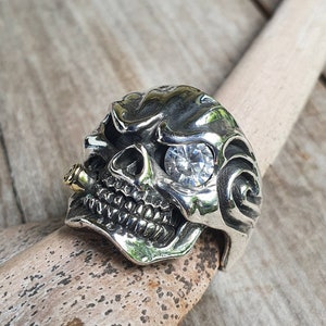 May include: A silver skull ring with a large, clear gemstone eye and a gold accent on the side. The ring is detailed with intricate carvings and sits on a wooden surface.