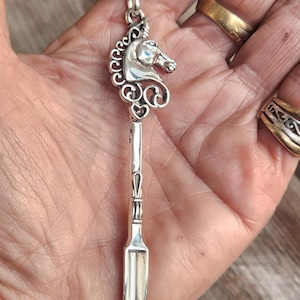 May include: A silver pendant necklace with a seahorse design. The pendant features a detailed seahorse head and a long, spoon-like body. The necklace is shown against a natural wood background.