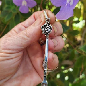 May include: A silver-colored pendant with a rose design at the top and a small scoop at the bottom. The pendant is held in front of a background of purple flowers and green foliage.