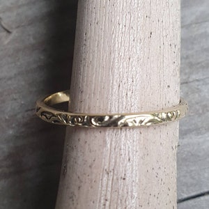May include: A gold-colored ring with an engraved floral pattern. The ring is displayed on a wooden dowel, showcasing its intricate design. The ring's band is thin, and the floral pattern adds a touch of elegance to the jewelry.
