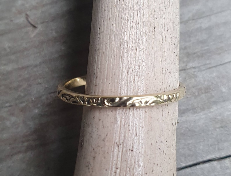 May include: A gold-colored ring with an engraved floral pattern. The ring is displayed on a wooden dowel, showcasing its intricate design. The ring's band is thin, and the floral pattern adds a touch of elegance to the jewelry.