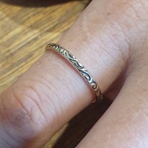 May include: A gold-colored ring with an intricate, repeating pattern of swirling lines. The ring is worn on a finger, showcasing the detailed design. The ring's surface reflects light, highlighting the texture and craftsmanship.