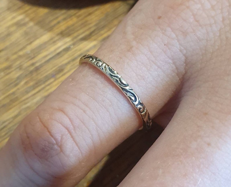 May include: A gold-colored ring with an intricate, repeating pattern of swirling lines. The ring is worn on a finger, showcasing the detailed design. The ring's surface reflects light, highlighting the texture and craftsmanship.