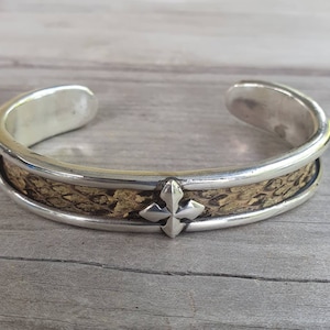 Könnte beinhalten: Silbernes Armreif-Armband mit einem strukturierten goldfarbenen Mittelband und einem silbernen kreuzförmigen Akzent. Das Armband hat eine polierte Oberfläche und ein schlichtes, offenes Design. Der Schmuck wird auf einer Holzoberfläche präsentiert.