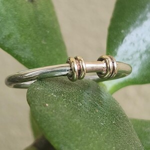 May include: A silver ring featuring two gold-coloured bands and a central gold-coloured bar. The ring is set against a blurred green leaf background, emphasising the jewellery's design and materials.