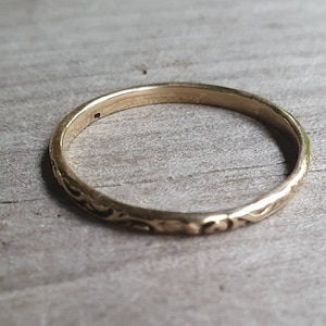 May include: A gold-colored ring with an engraved floral pattern. The ring is thin and appears to be made of metal. The ring is set against a light-colored wooden surface.
