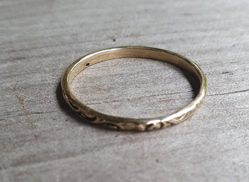 May include: A gold-colored ring with an engraved floral pattern. The ring is thin and appears to be made of metal. The ring is set against a light-colored wooden surface.