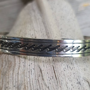 Könnte beinhalten: Ein silbernes Armreif-Armband mit einem Wellenmuster. Das Armband besteht aus poliertem Metall und weist ein detailliertes, geätztes Wellenmuster auf, das sich um die Mitte des Bandes legt. Das Armband ist etwa 1,9 cm breit.