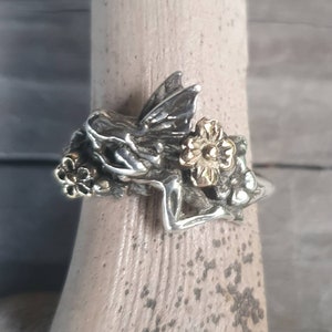 May include: A silver ring with a fairy design and gold-coloured flower accents. The fairy has detailed wings and is surrounded by floral elements. The ring is displayed on a wooden surface.