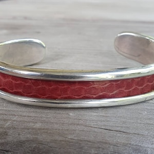 Könnte beinhalten: Ein silbernes Armreif-Armband mit einem roten, strukturierten Band. Das Armband hat eine polierte Silberoberfläche und ein breites, offenes Design. Das rote Band scheint aus Leder oder einem ähnlichen Material zu bestehen.
