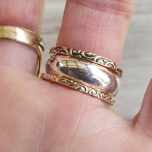 May include: Close-up of two rings. One is a wide, gold band. The other is a two-tone ring with a silver center and gold filigree edges. The rings are on a finger, with a blurred background.