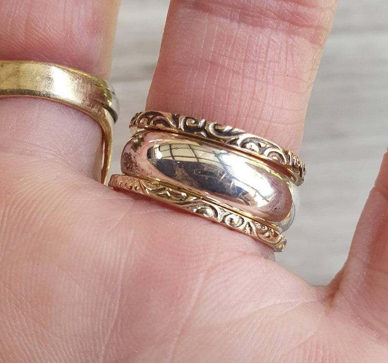 May include: Close-up of two rings. One is a wide, gold band. The other is a two-tone ring with a silver center and gold filigree edges. The rings are on a finger, with a blurred background.