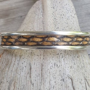 Könnte beinhalten: Ein silbernes Armreif-Armband mit einem Mittelband mit einem strukturierten, schuppenartigen Muster in Braun- und Beigetönen. Das Armband hat eine polierte, metallische Oberfläche und wird auf einer hellen Holzoberfläche präsentiert.