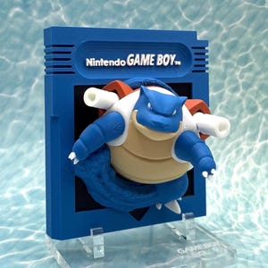 Könnte beinhalten: Eine blaue Skulptur in Form einer Nintendo Game Boy-Cartridge, die ein 3D-Modell des Pokémon Blastoise zeigt. Die Skulptur hat einen blauen Rahmen mit dem Text "Nintendo GAME BOY" und einen transparenten Ständer.