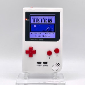Op de afbeelding: Een witte Game Boy Pocket draagbare videogameconsole met een rode en zwarte knopindeling. Het scherm toont het spel Tetris met de tekst "TETRIS TM" en "©1989 Nintendo".
