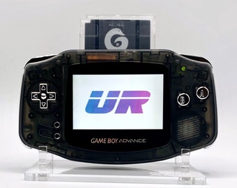 May include: A black Game Boy Advance handheld gaming console with a clear plastic shell. The console is displaying the logo for the game "UR" on the screen. A game cartridge is inserted into the console.
