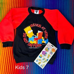 May include: Black and red long-sleeve sweatshirt with a cartoon image of Bart Simpson from The Simpsons. The sweatshirt has the text "Genius at Work Do Not Disturb" and a sticker with the text "The Simpsons Puffy Stickers".