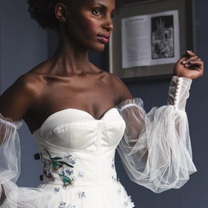 Op de afbeelding: Een witte off-shoulder bruidsjurk met een getailleerd lijfje en een volle rok. Het lijfje is versierd met geborduurde bloemen en de jurk heeft doorschijnende, gepofte mouwen.