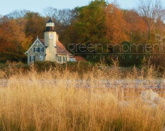 May include: A white lighthouse with a black roof stands in front of a backdrop of fall foliage. The trees are a mix of orange, yellow, and green. The lighthouse is surrounded by tall, golden grass.