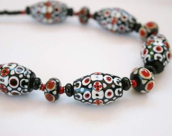 May include: A black and white necklace with red accents. The necklace is made of oval-shaped beads with a geometric pattern.