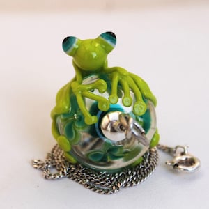 Könnte beinhalten: Ein grünes Glasfrosch-Figürchen mit blauen Augen sitzt auf einem Glas-Kugelanhänger. Der Anhänger hat einen silberfarbenen Schlüssel und ist an einer silbernen Kette befestigt. Der Frosch ist leuchtend grün.