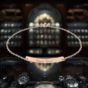 Può includere: Un braccialetto in oro rosa con catena e barra curva. La barra è incisa con la scritta "Your Text". Il braccialetto è esposto in una gioielleria, con orologi sullo sfondo.