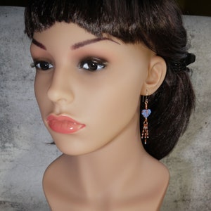 May include: A dangle earring featuring a light blue carved leaf bead suspended from a copper wire hook. Below the leaf, three thin copper wire strands are adorned with small, multi-colored seed beads in shades of brown, white, and blue.
