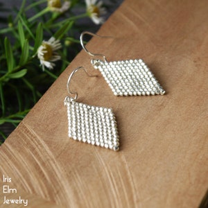 May include: Silver-toned, diamond-shaped earrings. Each earring is crafted from small, closely-set beads, creating a textured surface. The earrings hang from curved ear wires. The image also shows the text "Iris Elm Jewelry".