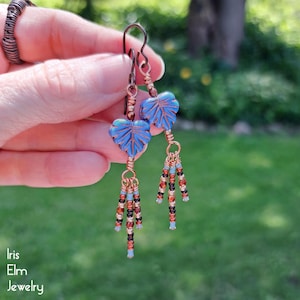 May include: Dangle earrings featuring blue leaf-shaped beads with copper wire wrapping and dangling fringe made of small metallic copper, black, and light blue seed beads. The earrings hang from dark copper-toned metal ear wires.