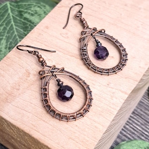 May include: A pair of teardrop-shaped earrings crafted from oxidized copper wire, featuring intricate wire-wrapped detailing and a single faceted, deep purple bead suspended in the center of each frame, complete with matching copper ear hooks.