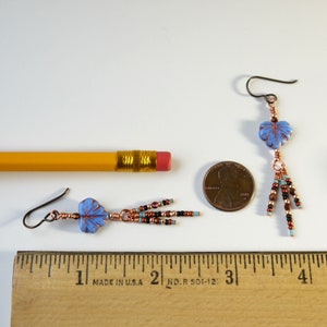 May include: A pair of dangle earrings featuring light blue leaf-shaped beads with copper wire wrapping. Each earring has three beaded fringe strands in brown, black, and light blue. A yellow pencil and a US penny provide scale reference.