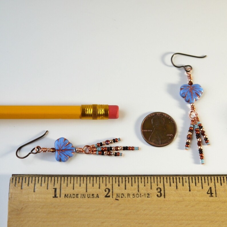 May include: A pair of dangle earrings featuring light blue leaf-shaped beads with copper wire wrapping. Each earring has three beaded fringe strands in brown, black, and light blue. A yellow pencil and a US penny provide scale reference.